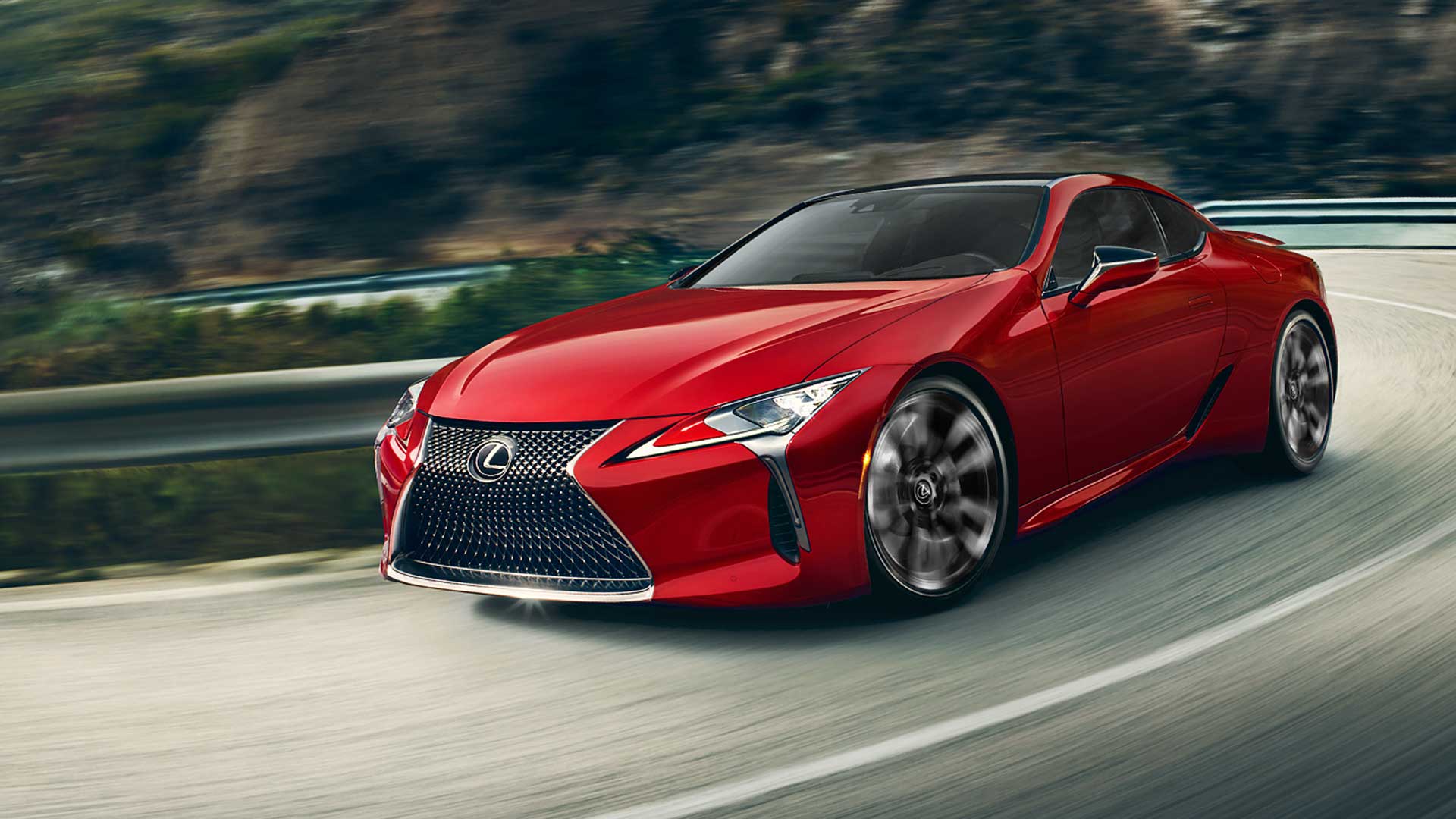 A red LC 500 Coupe navigates a winding road. There is blurred greenery in the background.