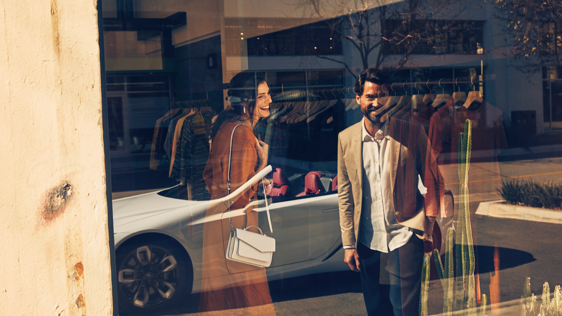 The reflection of an LC Convertible and a smiling man and woman are seen in a shop window.